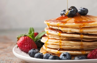 Golden pancake syrup being poured over a stack of pancakes with fresh blueberries and strawberries